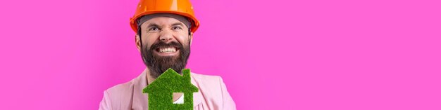 Portrait of young construction engineer wear orange hard hat in a pink jacket standing on red studio background A man holds a green eco house