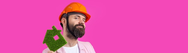 Portrait of young construction engineer wear orange hard hat in a pink jacket standing on red studio background A man holds a green eco house