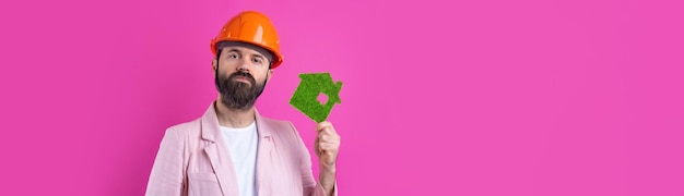 Portrait of young construction engineer wear orange hard hat in a pink jacket standing on red studio background A man holds a green eco house