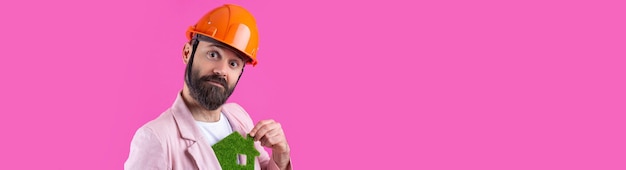 Portrait of young construction engineer wear orange hard hat in a pink jacket standing on red studio background A man holds a green eco house