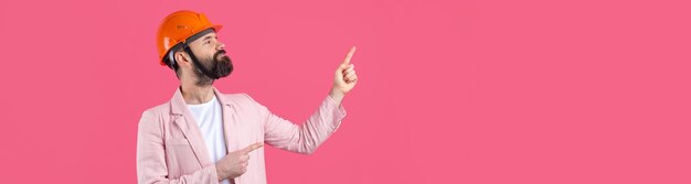 Portrait of young construction engineer wear orange hard hat in a pink jacket standing on red studio background The man points with his hand