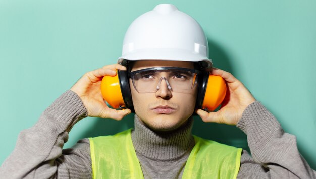 Foto ritratto di giovane operaio ingegnere edile che indossa il casco di sicurezza, occhiali di protezione, giacca e cuffie su sfondo di colore aqua menthe.