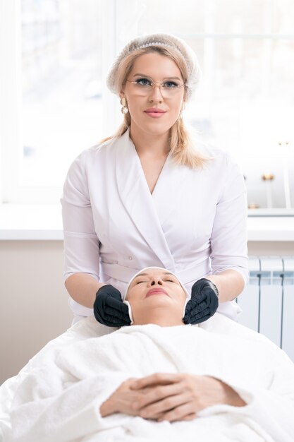 Ritratto di giovane cosmetologo in guanti neri utilizzando un batuffolo di cotone durante la pulizia della pelle del viso di donna matura prima della procedura