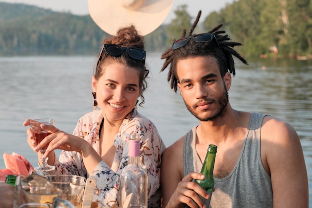 Portrait of young couple drinking beer and smiling at camera while sitting at the table during lunch outdoors