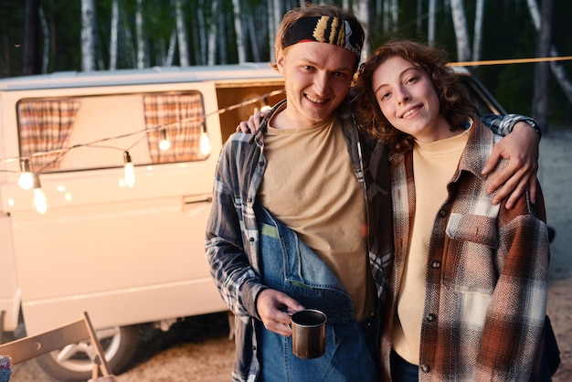 Portrait of young couple embracing and smiling at camera while standing in the forest during camping