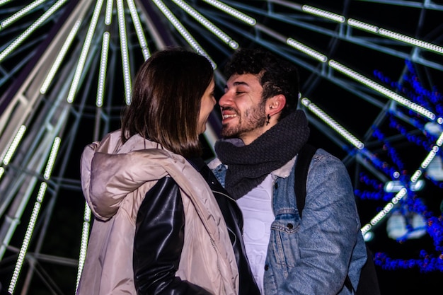 クリスマステーマパークの観覧車の前でキスする若いカップルのポートレート