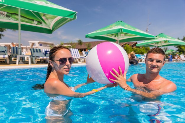 Ritratto di giovane coppia in piscina che tiene insieme il pallone da spiaggia a strisce e che guarda l'obbiettivo - testa e spalle delle coppie in vacanza e divertirsi nella piscina del resort