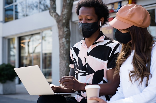 Portrait of young couple wearing protective mask and using laptop outdoors on the street