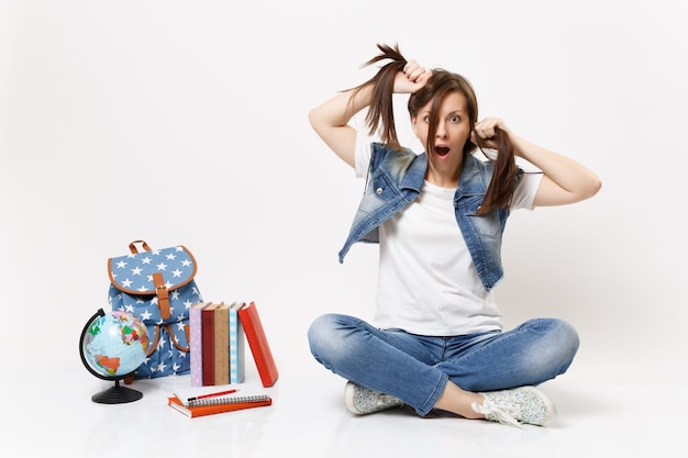 Portrait of young crazy shocked woman student in denim clothes holding ponytails, sitting near globe, backpack, school books isolated