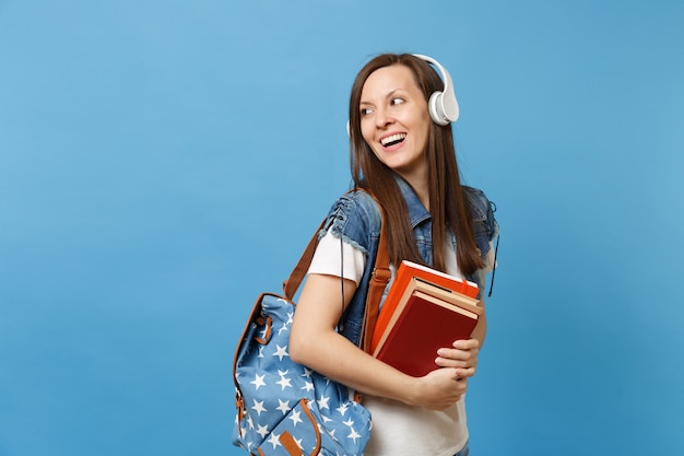 Portrait of young curious woman student with backpack headphones listening music hold school books looking aside on copy space isolated on blue background. Education in high school university college.