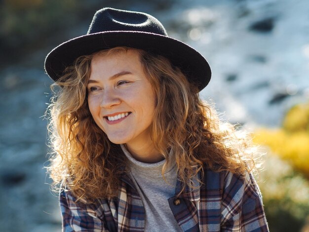 Foto ritratto di una giovane donna riccia con cappello di feltro e maglietta a quadri con fiume di montagna