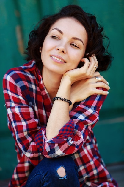 Ritratto di giovane donna riccia sulla parete verde. ragazza sorridente ed emozionale in jeans e camicia rossa sulla strada della città. giovane donna sveglia all'aperto nel giorno soleggiato