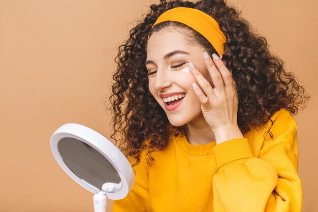 Portrait of young curly woman using skin cream on in a mirror isolated over beige background. Skincare concept.