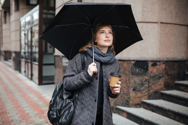 黒い傘とコーヒーを手に通りに立っている若いかわいい女性の肖像画