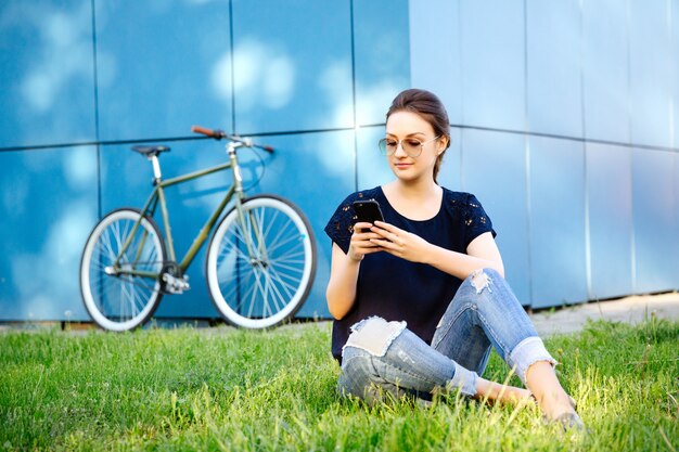 Portrait of young cute woman with smartphone, browsing something or chatting while sitting on grass, after riding a bike. Leisure time.