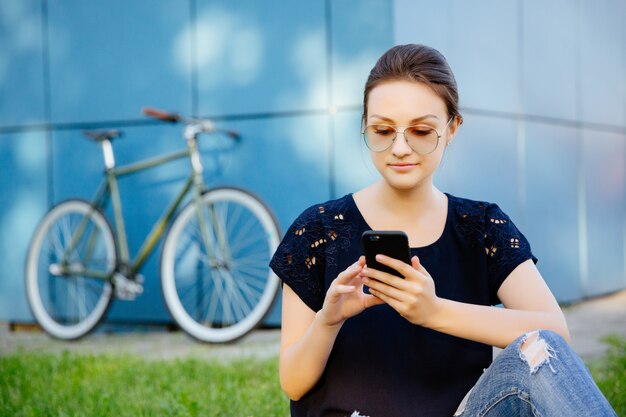 Portrait of young cute woman with smartphone, browsing something or chatting while sitting on grass, after riding a bike. Leisure time.