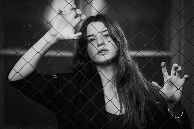 Портрет Молодая темноволосая женщина, которая стоит за сетчатым забором на расстоянии Концепция одинокой и грустной женщины