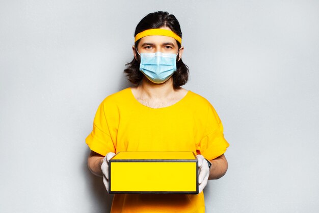 Portrait of young delivery man holding a yellow box. Wearing medical mask, surgical gloves and orange shirt. Background of grey textured wall.