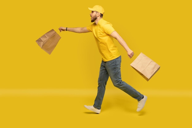 Portrait of young delivery man in yellow uniform running with paper packets isolated over yellow background