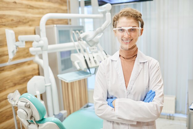 Portrait of young dentist in white coat and in protective glasses standing with arms crossed and smiling at front in clinic