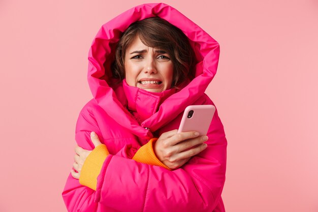 Portrait of young displeased woman in warm coat is freezing and using cellphone isolated on pink