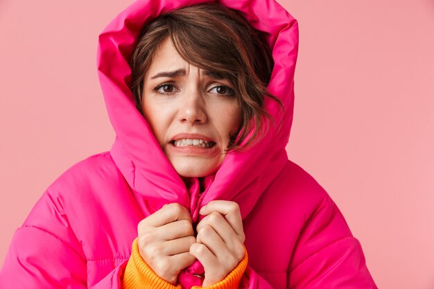 Portrait of young displeased woman in warm hood is freezing isolated on pink