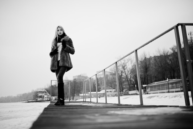 Portrait of young elegance blonde girl in a fur coat at pier background foggy river on winter ice