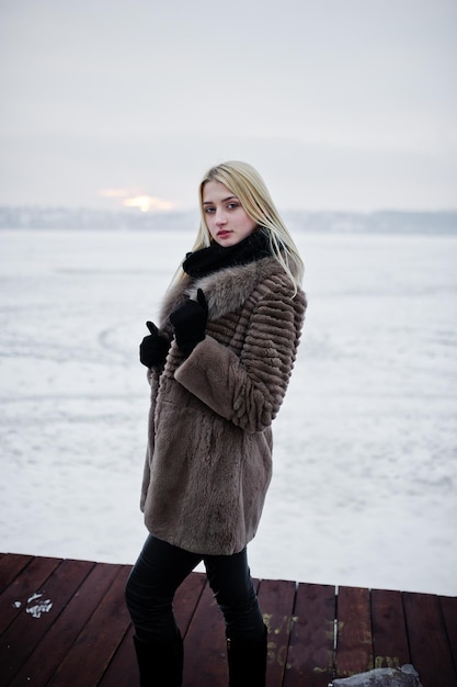 Portrait of young elegance blonde girl in a fur coat at pier background foggy river on winter ice