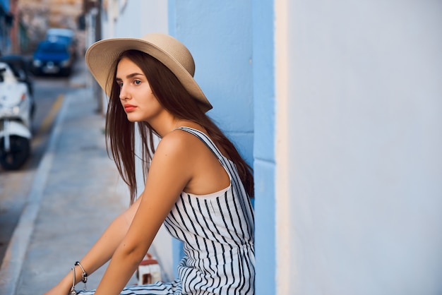 Ritratto di giovane donna elegante in cappello di paglia seduto sulla strada.