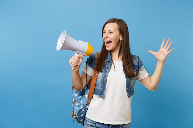 バックパックとデニムの服を着た若い感情的な女性の学生の肖像画は、青い背景で隔離の手を広げて叫んでメガホンを保持します。高校での教育。広告用のスペースをコピーします。