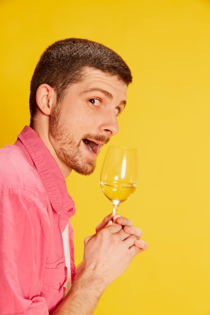 Portrait of young emotive man man in pink shirt posing with glass of white wine isolated over vivid yellow background