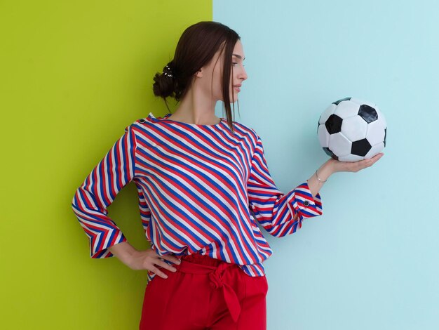 Portrait of young European woman holding soccer ball on her palm. Happy girl, football fan or player isolated on green and blue background. Sport, play football, health, healthy lifestyle concept