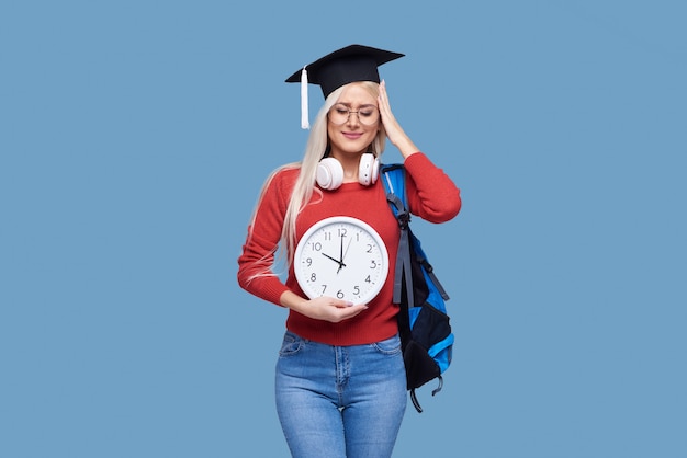 灰色の空間に分離された大きな目覚まし時計を保持しているバックパックと大学院のキャップで若い興奮した金髪の女性学生の肖像画。大学での教育。テキストのスペースをコピー