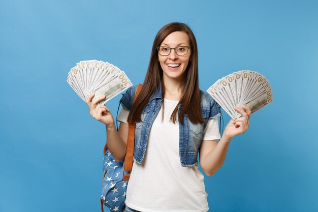 Portrait of young excited joyful woman student in glasses with backpack holding bundle lots of dollars, cash money isolated on blue background. Education in high school university college concept.