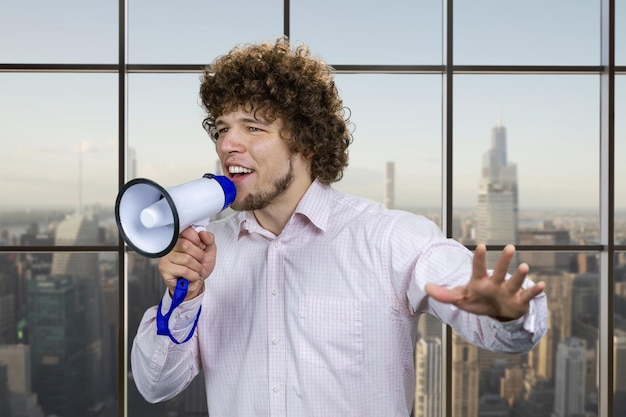 Photo portrait of a young excited man with curly hair in white shirt screaming hot news shout in megaphone