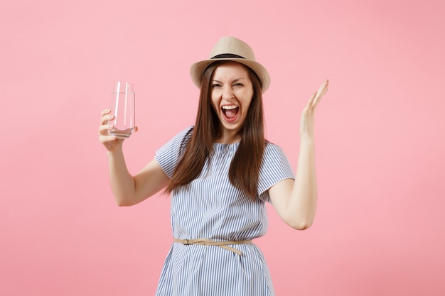 Portrait of young excited woman in blue dress, hat holding and drinking clear fresh pure water from glass isolated on pink background. Healthy lifestyle, people, sincere emotions concept. Copy space.