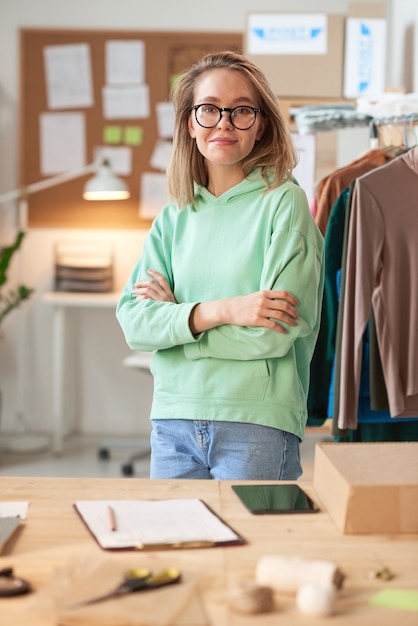 Portrait of young fashion designer in eyeglasses standing with arms crossed and looking at front during her work at office