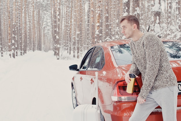 Portrait of young fashionable man in grey clothes with car on snow holding suitcase and go pathway pine forest. Winter vacation travel concept. Outdoors in winter. Ski resort, mounting skiing resort