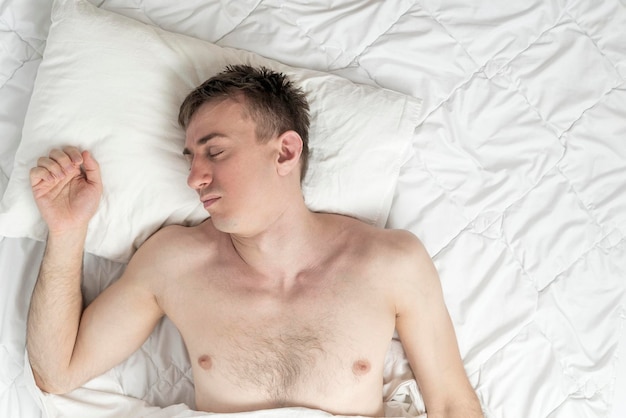 Портрет молодого быстро спящего мужчины, вид сверху. Здоровый сон. Парень спит на белом хлопковом постельном белье.