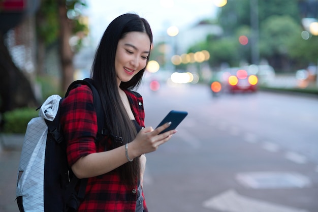 Portrait of young female backpacker standing near the street using smartphone waiting for taxi