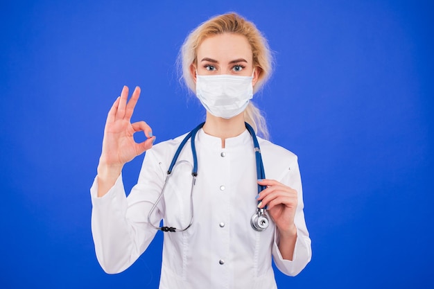 Portrait of a young female doctor in a protective mask shows the OK gesture on a blue background
