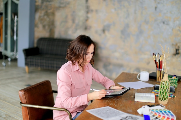 Ritratto di giovane libero professionista femminile che utilizza la compressa del touch screen per il lavoro a distanza mentre sedendosi nell'interno moderno