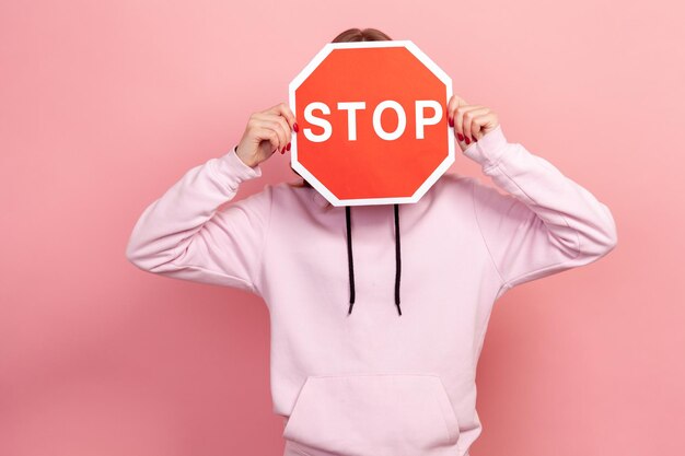 Portrait of young female in hoodie covering face with red road stop sign warning to go prohibition concept Indoor studio shot isolated on pink background