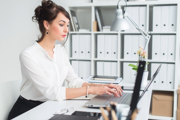 Portrait of a young female office worker sitting at her workplace typing, looking on laptop screen attentively.