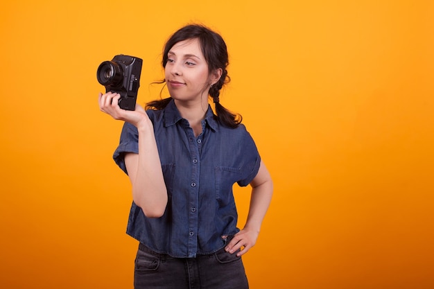 Portrait of young female photographer holding her camera in studio over yellow background. Beautiful caucasian girl looking at her photo camera.