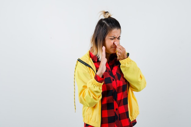 Portrait of young female pinching nose due to bad smell, showing palm in checkered shirt, jacket and looking disgusted front view