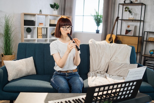 Portrait of young female playing on flute Pretty girl learning to play classic flute at home