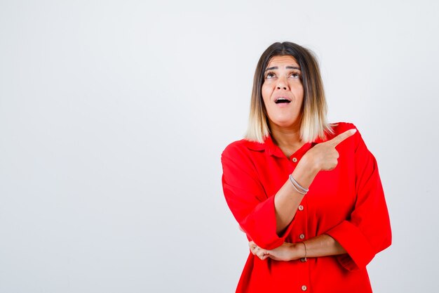 Portrait of young female pointing at upper right corner in red oversized shirt and looking puzzled front view