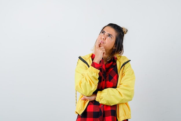 Photo portrait of young female standing in thinking pose, looking up in checkered shirt, jacket and looking puzzled front view