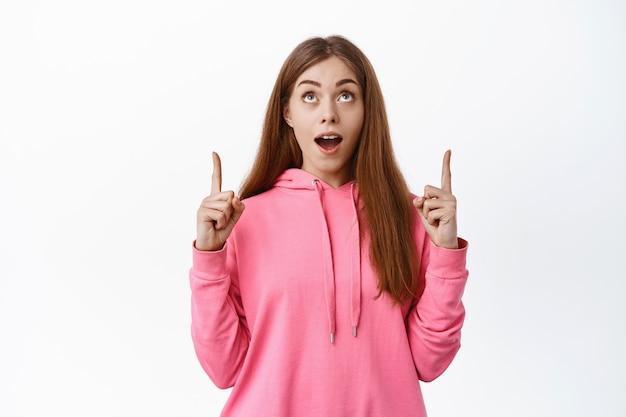 Portrait of young female student looks impressed and points fingers up, shows advertisement, stands over white wall